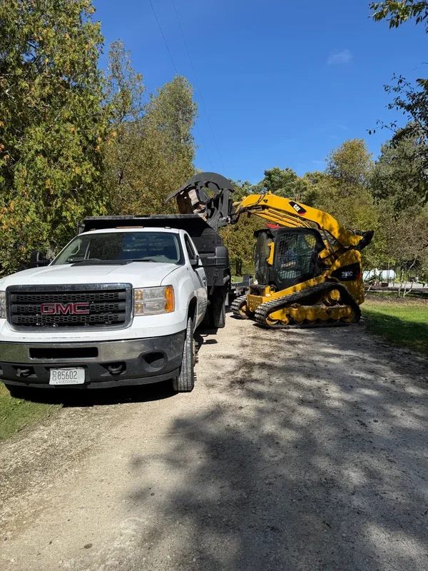 Skid steer and dump truck working along a shaded gravel road