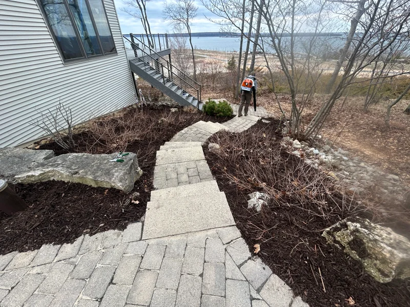 Crew member blowing debris from stone steps beside mulched beds at a lakeside home